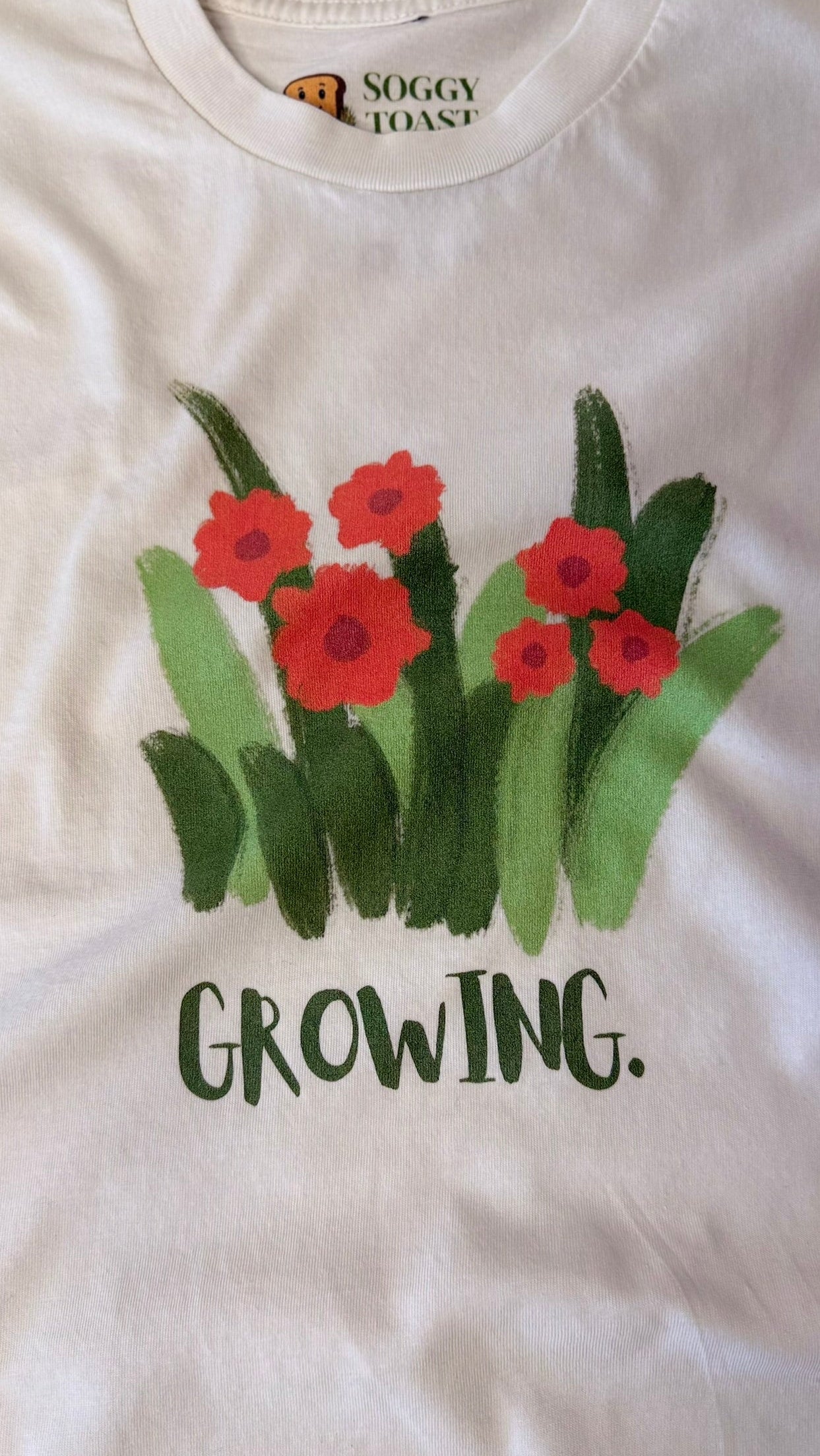 White t-shirt with red flowers and green leaves and the text 'GROWING.'