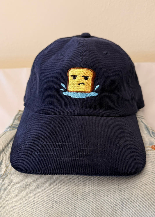 Navy cap with an embroidered design of a bread character in water on a beige background.